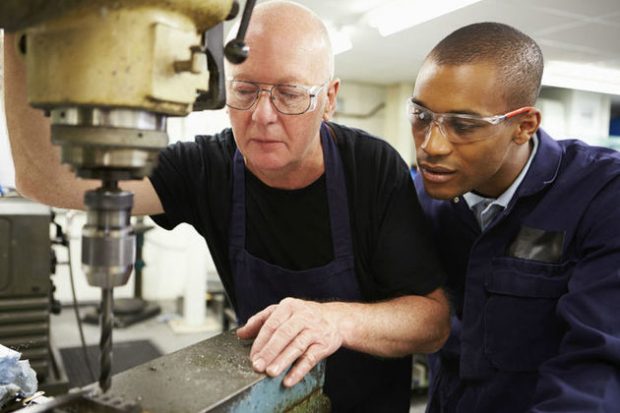 A young trainee wearing overalls and safety goggles observing an older colleague operating machinery