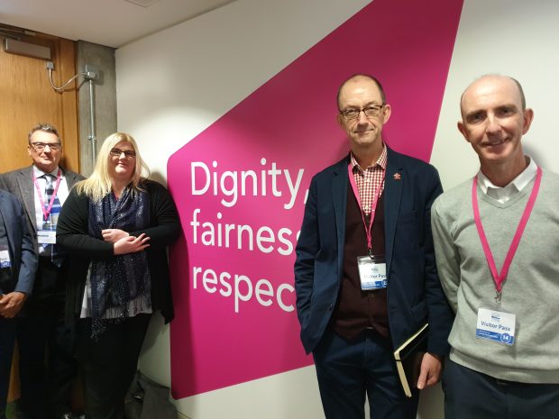 Social Security Advisory Committee members Bruce Calderwood, Victoria Todd, Phil Jones and Jim McCormick standing by a sign which says dignity, fairness and respect. Photograph taken at Social Security Scotland in Dundee.